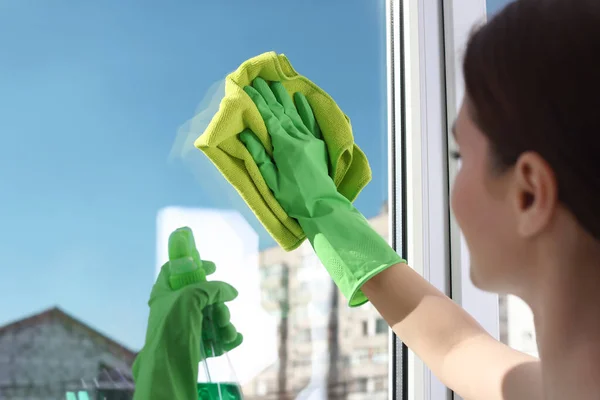 Side view of woman cleaning window glass at home with cloth and spray bottle close up