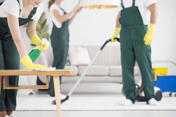 A team of cleaners in green overalls cleaning a room by vacuuming and dusting.